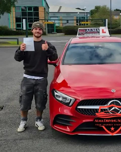 Learner holding a practical driving test pass certificate beside the Alma Driving School red Mercedes A-Class