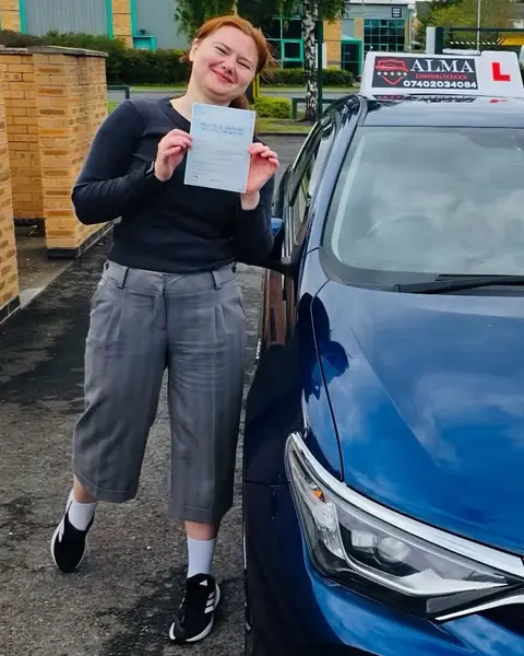 Learner holding a practical driving test pass certificate beside the Alma Driving School blue Toyota Auris