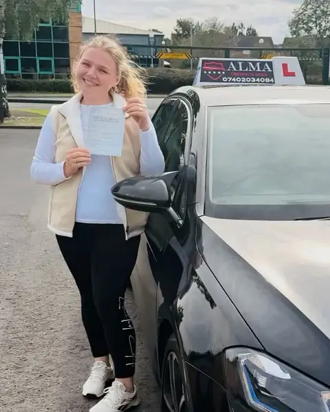 Learner holding a practical driving test pass certificate beside the Alma Driving School black VW Golf