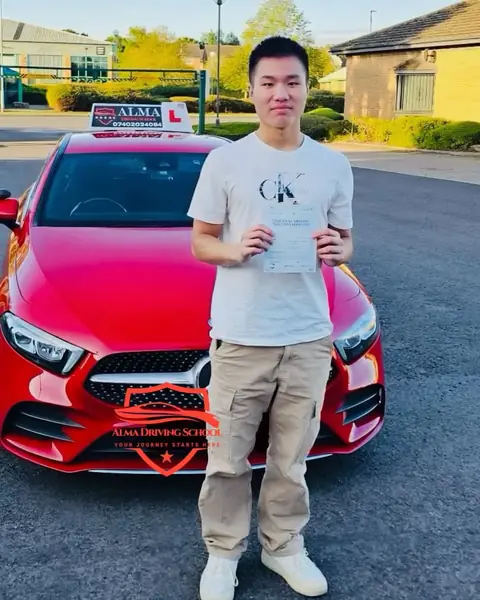 Learner holding a practical driving test pass certificate beside the Alma Driving School red Mercedes A-Class