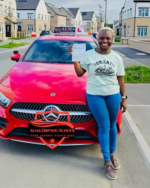 Learner holding a practical driving test pass certificate beside the Alma Driving School red Mercedes A-Class