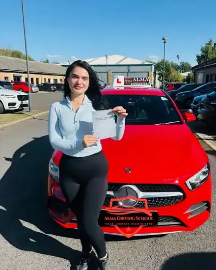 Learner holding a practical driving test pass certificate beside the Alma Driving School red Mercedes A-Class
