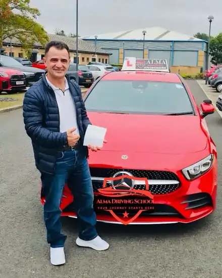 Learner holding a practical driving test pass certificate beside the Alma Driving School red Mercedes A-Class
