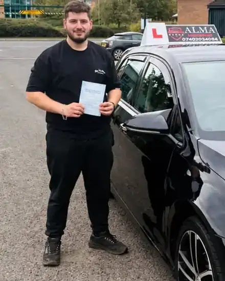 Learner holding a practical driving test pass certificate beside the Alma Driving School black VW Golf