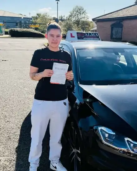 Learner holding a practical driving test pass certificate beside the Alma Driving School black VW Golf