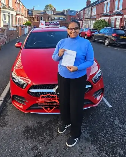 Learner holding a practical driving test pass certificate beside the Alma Driving School red Mercedes A-Class