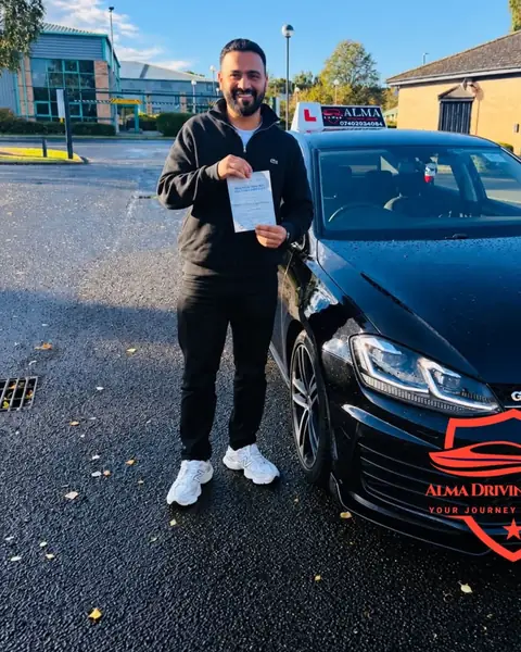 Learner holding a practical driving test pass certificate beside the Alma Driving School black VW Golf