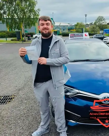 Learner holding a practical driving test pass certificate beside the Alma Driving School blue Toyota Auris