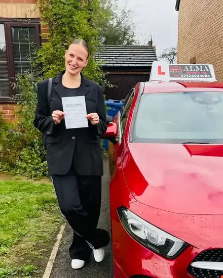 Learner holding a practical driving test pass certificate beside the Alma Driving School red Mercedes A-Class